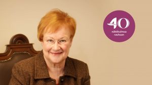 Presidentti Tarja Halonen hymyilee kameralle. Kuvassa on purppura logo, jossa lukee 40 Näkökulmaa rauhaan.