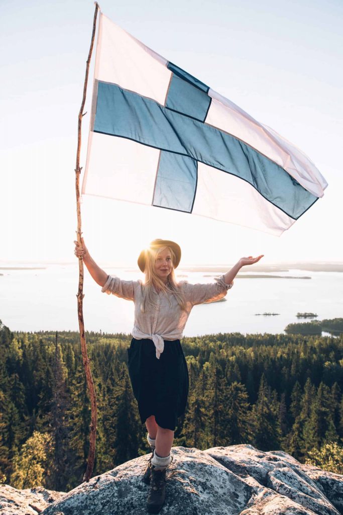 Saariselän kansallismaisemassa auringonnousussa seisoo nuori nainen joka pitää isoa Suomen lippua. Lipun salko on pitkä männynoksa. Hänen takanaan aukeaa metsämaisema ja järvimaisema, jossa on saaria.
