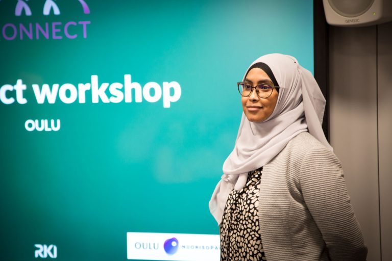 An image of Faiza Abdulqadir standing with her hands behind her back and a smile on her face. She is wearing glasses, a black and white pattern shirt, a light-coloured cardigan and a white scarf covering her hair. In the background there is a turquoise wall with the Connect logo on it.