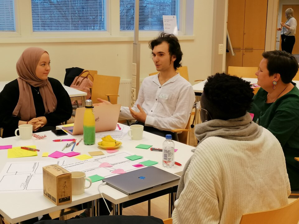 Pöydän ääressä istuu neljä ihmistä keskustellen. Pöydällä on mm. värikkäitä post-it lappuja, läppäri ja kahvikuppeja.